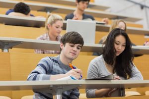 Students sitting in a lecture hall. Some students are taking written notes. One student is working on her laptop.